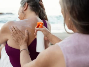 Person applying spray sunscreen to another's back at the beach